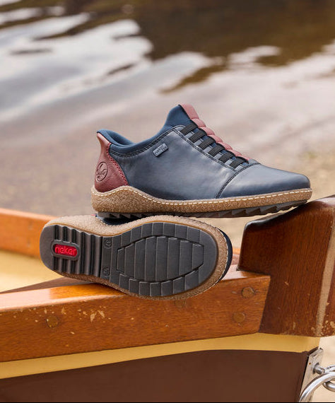 A single dark blue leather shoe with red accents and a brown sole, resting on the wooden edge of a boat with water and sand in the background.