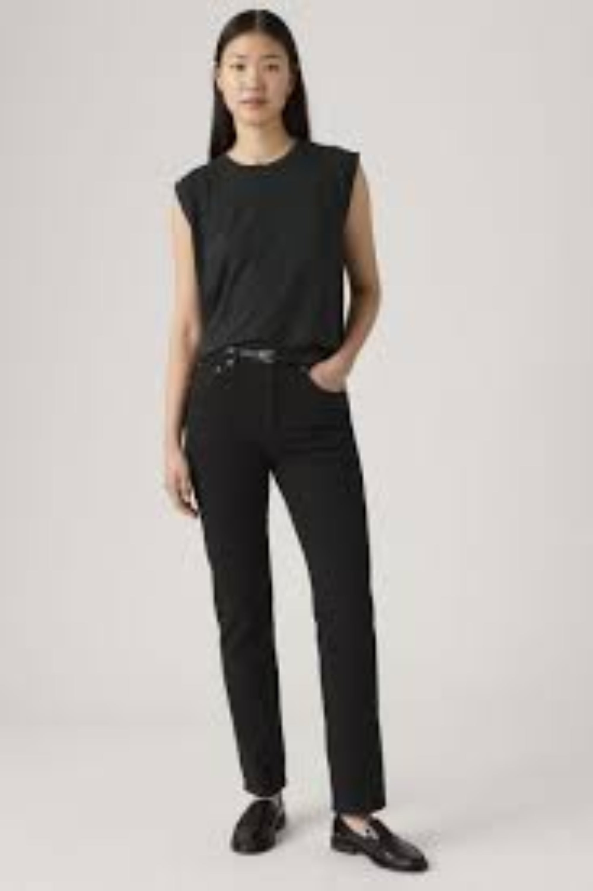 A model wearing a black sleeveless top and black pants with a thin black belt, standing against a light gray studio background.