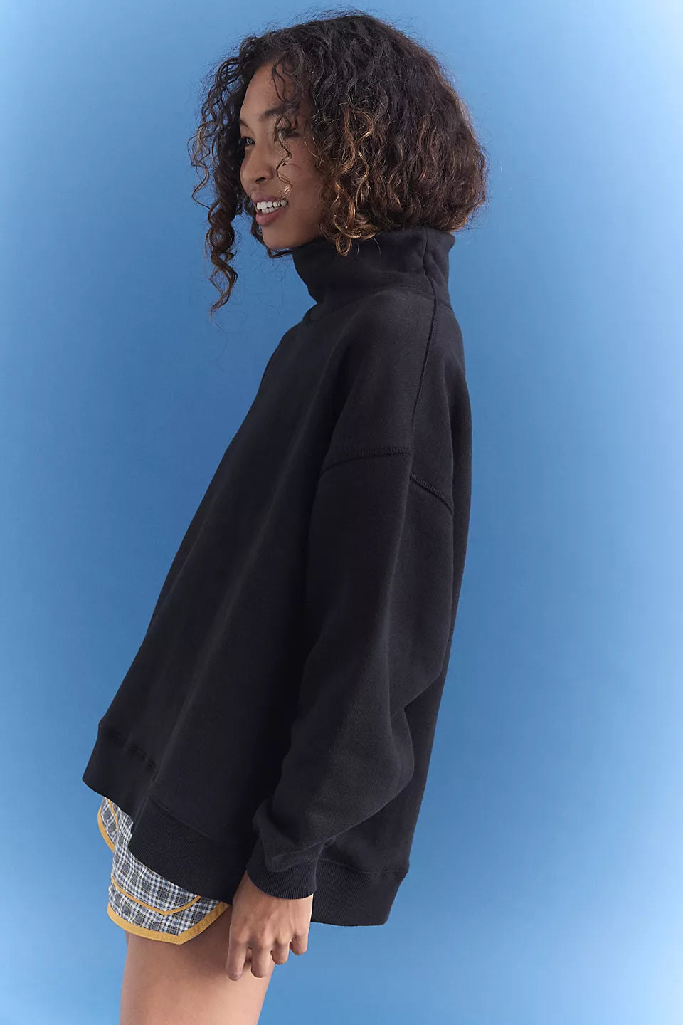 A woman with curly brown hair is shown in profile wearing an oversized black mock-neck sweatshirt and plaid shorts against a blue gradient background.