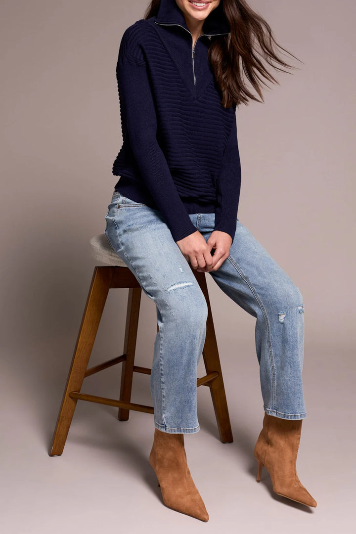 Person wearing a navy sweater, light blue jeans, and brown boots sitting on a wooden stool.