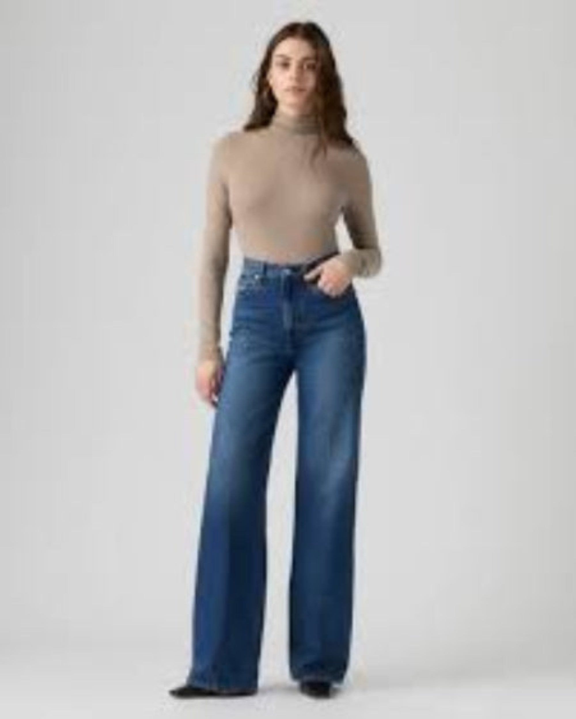 A woman modeling a beige mock-neck long-sleeve top and high-waisted, wide-leg blue jeans against a plain light gray studio background.