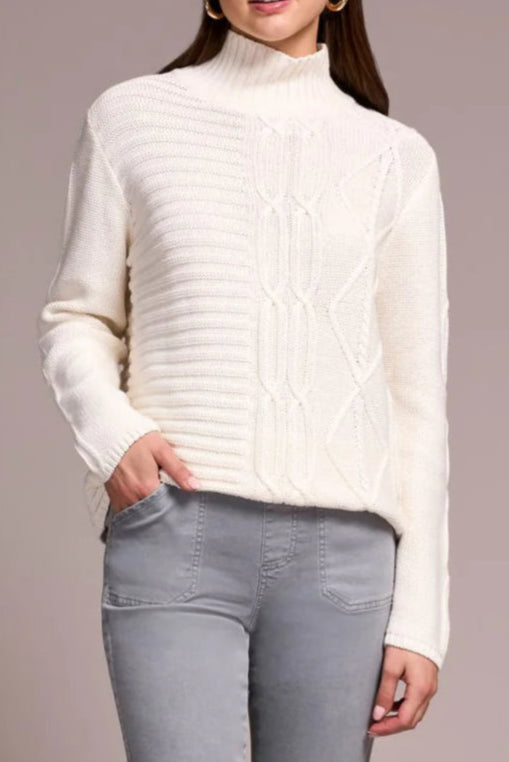 Woman wearing a white turtleneck sweater and gray jeans against a plain background