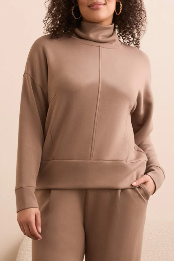 Woman wearing a beige tracksuit standing in a room with a plant and sofa.